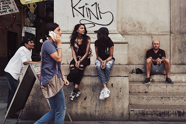 Naples, Italy – July 2017: A candid street scene on Via Toledo shows a group of people relaxing near an old building. Two young women sit on a step chatting, a man sits alone on the steps, and a person passes by talking on the phone. The image captures the lively, authentic, and multicultural atmosphere of Neapolitan urban life on a summer afternoon.