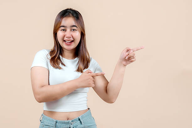 istock A cheerful young woman smiling widely while playfully pointing in both directions, set against a soft beige background, radiating joy and enthusiasm. 2242581381