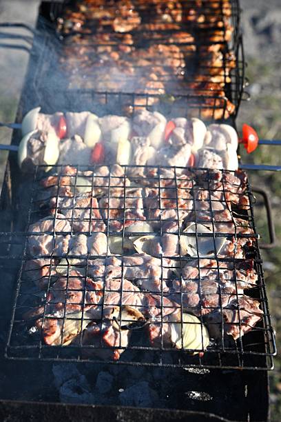 Vertical shot of a charcoal grill loaded with skewers and grill racks, actively cooking meat. Ideal for news articles, blogs, and reports on outdoor traditions, events, and picnic area usage.