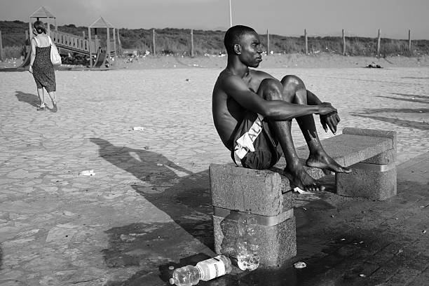 Castel Volturno, Italy – August 2017: An immigrant man sits on a concrete bench, facing the sea, which is not visible in the image. The black-and-white photograph captures a moment of silence and reflection, a symbol of solitude, dignity, and hope, against the backdrop of a sunny beach on the Campania coast.