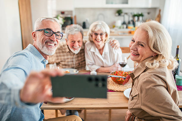 istock Two cheerful senior couples sitting around a dining table at home, enjoying lunch together. One senior man is holding a smartphone, taking a selfie photo of the group 2242593451
