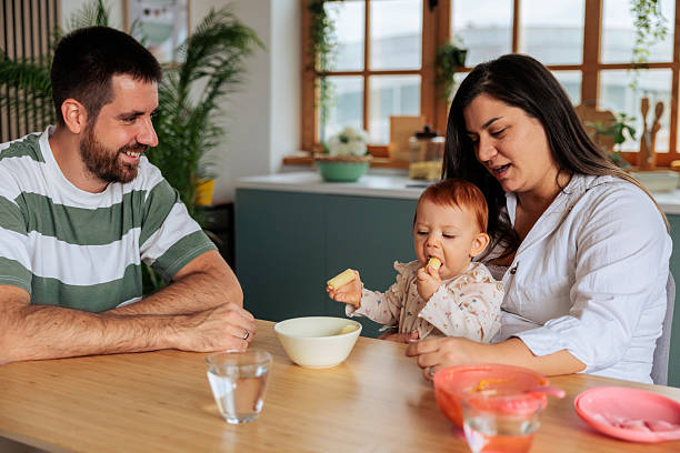 A happy young family is sharing a mealtime moment at their kitchen table. The parents smile warmly as the baby independently feeds herself a piece of food. This image beautifully captures the joyful experience of introducing solid foods and the bond during family life.