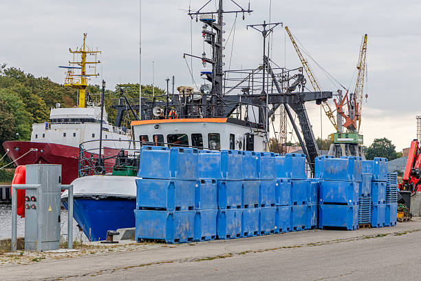 A busy industrial port scene featuring a fishing or service vessel docked, blue plastic containers stacked in the foreground, and yellow cargo cranes in the background under a gray sky.