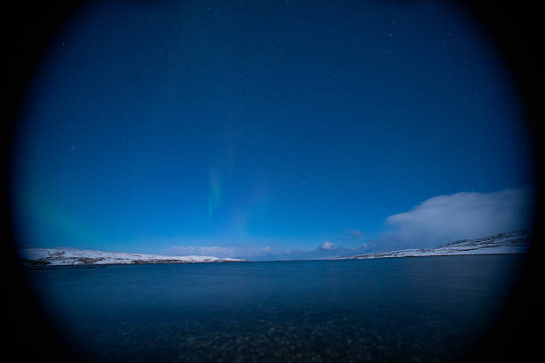 A night landscape featuring a faint vertical glow of the Northern Lights over a calm body of water with a rocky beach foreground and starry sky