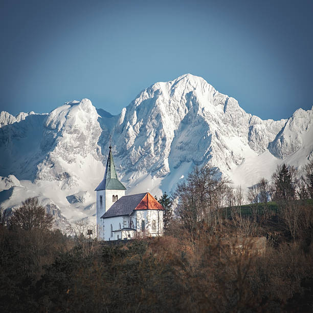 A picturesque white church with a red roof stands on a hill, with snow-capped mountains in the background under a clear blue sky.