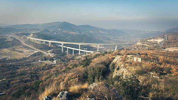 A long, white viaduct stretches across a valley, connecting two hillsides. The bridge is supported by numerous concrete pillars and has cars driving on it. The surrounding landscape is hilly and covered with trees, some of which have autumn foliage. The sky is clear and blue, with a slight haze in the distance.