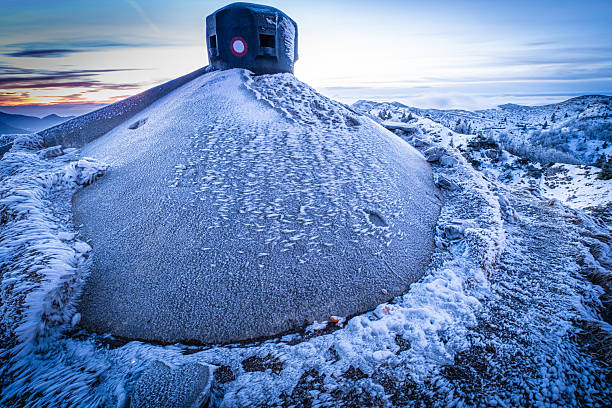 A concrete bunker, covered in frost and ice, sits atop a snow-covered mountain under a dramatic sky at sunrise. The bunker, likely a remnant of past conflicts, stands as a stark reminder of history against the backdrop of a serene, frozen landscape.