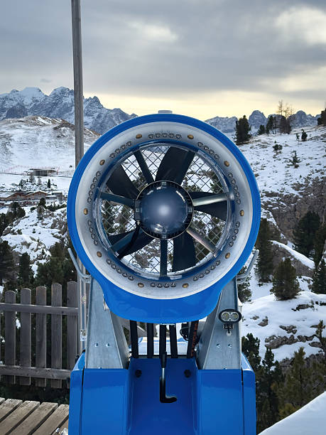 Close-up of a modern snow cannon on a snowy slope in the Dolomites, Italy, surrounded by alpine peaks and cloudy winter sky