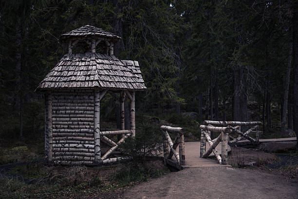 A dramatic, wide shot of a rustic, handmade wooden gazebo (shelter or small cabin) constructed from logs and a shingled roof, connected by a small wooden bridge. The scene is set in a dark, dense, and spooky conifer forest, conveying a sense of solitude, mystery, and an untouched, northern wilderness.