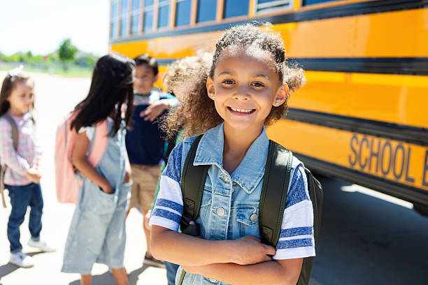 istock Happy young student smiles at the camera while waiting by a yellow school bus 2242651900