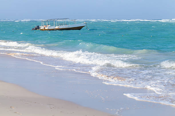 A small motor boat floats off a white sandy beach as turquoise waves roll in under a bright, clear sky. Calm seaside scene perfect for travel, vacation, and relaxation themes. Dominican Republic
