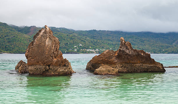 Battered rock formations emerge from calm turquoise sea, set against lush green mountains and a misty, overcast sky. A serene coastal landscape capturing nature, travel, and tropical scenery. Samana