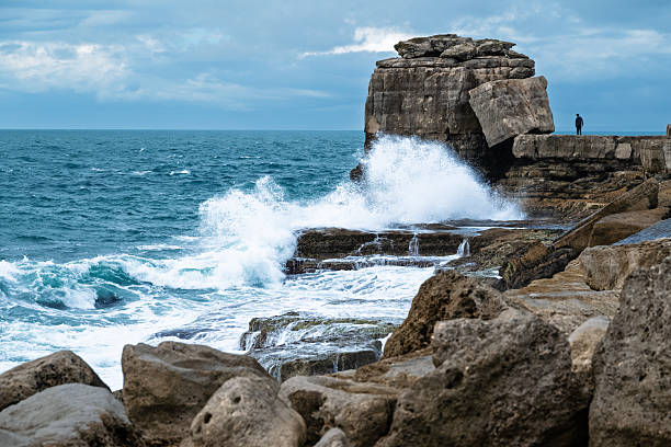 Dramatic waves crash against a rocky outcrop as a person stands on the cliff edge.
