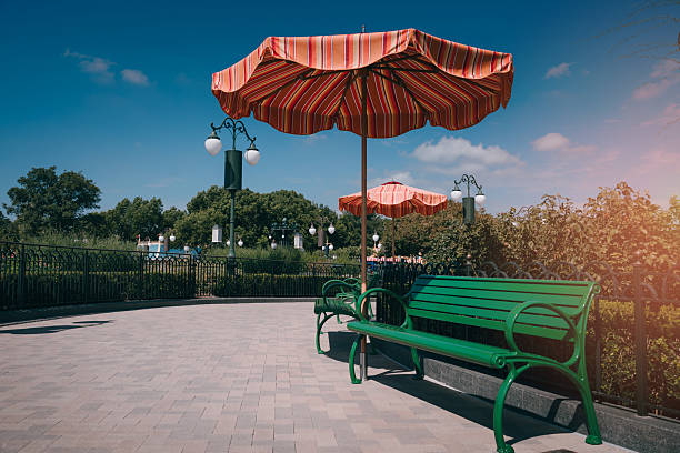 Colorful striped umbrellas casting shade over empty green benches in a tranquil park setting under a clear blue sky.