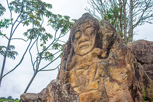 An ancient rock carving depicting a figure in San Agustin, Huila, Colombia, surrounded by lush greenery and cloudy skies.