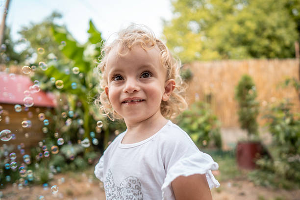 A happy young girl in a white shirt smiles at colorful bubbles in a sunlit backyard. Lush greenery and a wooden fence create a warm, playful outdoor mood.