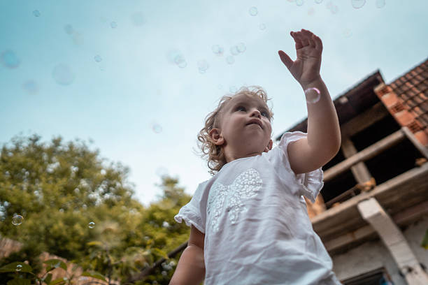 A young child reaches upward to touch bubbles fluttering in a bright outdoor setting. The scene combines playful innocence with a backdrop of trees and a partially built structure, conveying carefree outdoor fun.