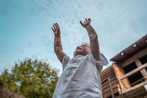 A joyful child reaches upward amid bubble spheres in a sunny outdoor setting, conveying play, wonder, and carefree mood. Perfect for themes of summer, family, and childhood joy.