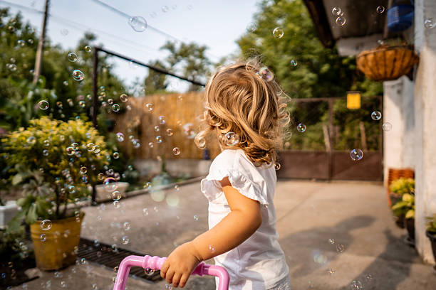 A joyful toddler girl in a white shirt pushes a pink scooter handle while bubbles float around in a sunny backyard. A warm, playful moment perfect for family and outdoor activity themes.