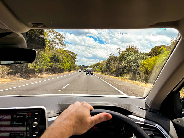 istock A wide-angle view through a car’s windscreen reveals a highway through a forested area, framed by the interior, steering wheel, and driver’s hands under a clear blue sky with fluffy clouds 2242678879