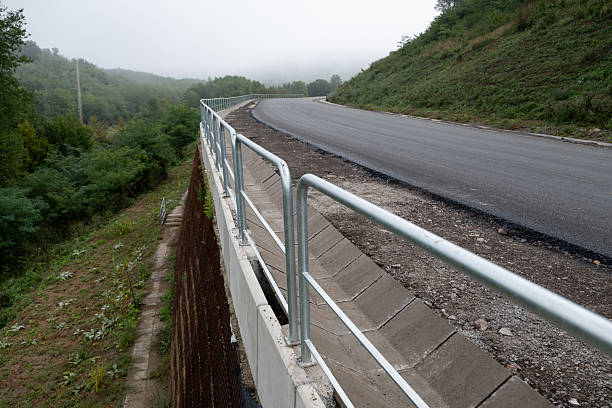 A freshly asphalted mountain road curves gently through a misty green landscape. A sturdy metal safety railing lines the edge of the road, ensuring protection along the elevated slope. The smooth surface and foggy atmosphere emphasize progress in infrastructure and harmony with nature.