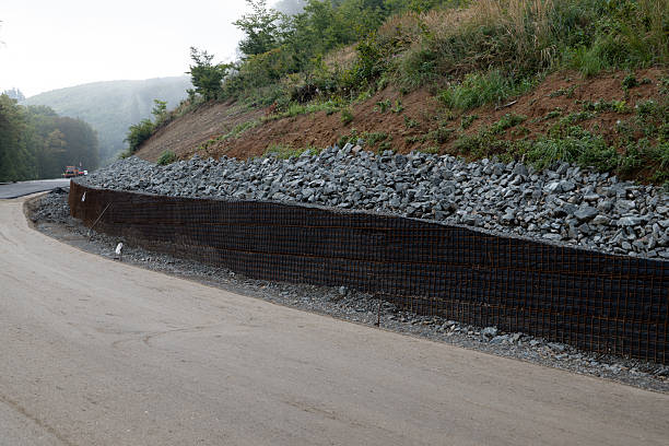 A reinforced retaining wall made of metal mesh and rocks lines a curved mountain road under construction. The structure stabilizes the slope and prevents erosion, ensuring the durability and safety of the new roadway. Misty hills and greenery surround the scene, blending engineering with nature.