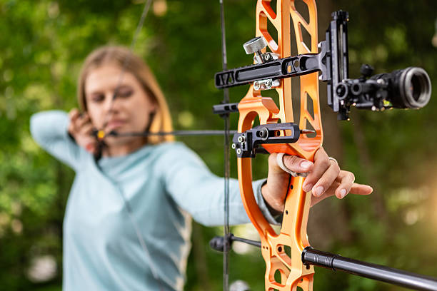 Zoung Caucasian female archer aiming her compound bow with concentration outdoors. She wears a light blue long-sleeved top, demonstrating precise focus in a natural setting. The orange bow and sight are prominently visible, highlighting her archery skills.