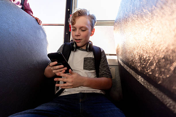 istock Young boy on bus using smartphone, headphones around his neck, sunlight in background 2242687353