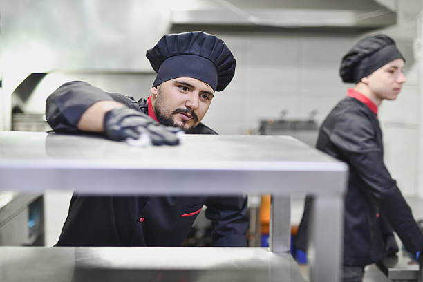 Two professional chefs work in a modern kitchen, one leaning over a metal surface, concentrating on a task. The scene conveys teamwork, skill, and the food service environment.