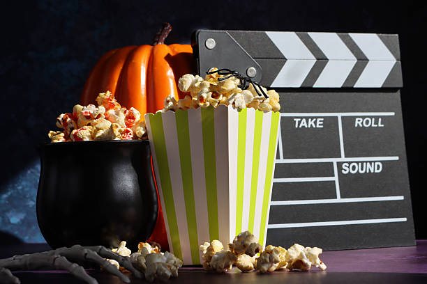 istock Image of plastic spider balancing on popcorn kernels in green and white striped popcorn carton, cauldron film snack food tub, film slate, skeletal hand, orange pumpkin background, focus on foreground, Halloween scary film concept 2242691749