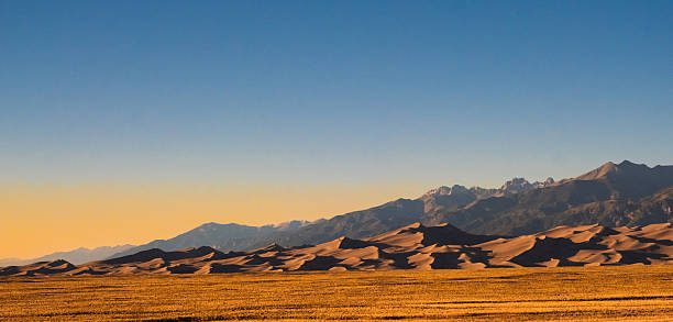 A simple yet breathtaking  sunset over the sweeping dunes of Great Sand Dunes National Park in Colorado. As the sun descends, warm hues of orange and gold flood the sky, casting long shadows across the rippled sand. The distant mountains seem to glow with the last light, their peaks gradually fading into twilight. As night approaches, the sky deepens to a rich indigo, completing the mesmerizing cycle of day turning into night amidst the vast, quiet dunes.
