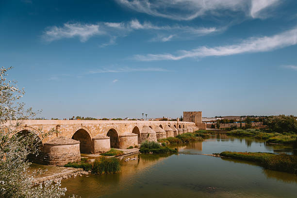 Ancient Roman bridge in Córdoba spanning the Guadalquivir River with repeating stone arches, clear blue sky and tranquil reflections showcasing historic Andalusian architecture
