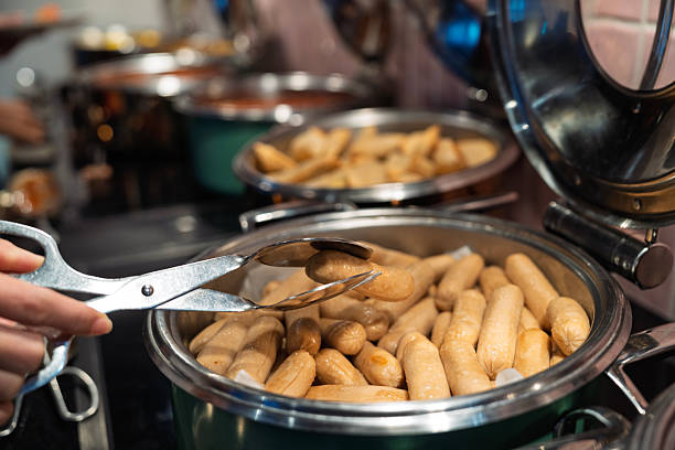 A close-up view of breakfast sausages in a stainless steel chafing dish at a buffet station, with a hand using tongs to pick up a piece.