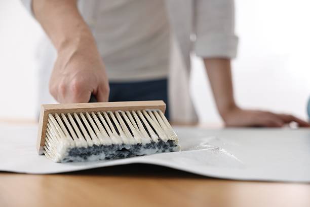 istock Man applying glue onto wallpaper sheet at table indoors, closeup 2242708102