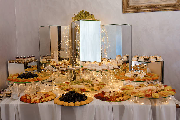 Luxurious dessert table setup featuring mirror columns, tiered platters laden with fruits and pastries, soft white linens, and warm decorative lighting for weddings and celebrations