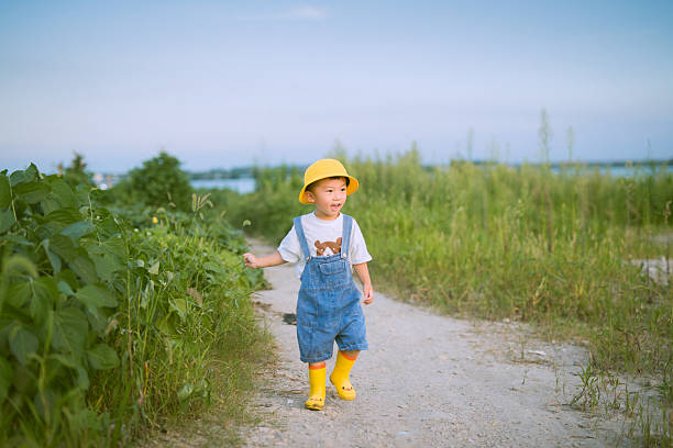 A cheerful young boy in a yellow bucket hat, blue overalls, and yellow rain boots walks along a dirt path surrounded by lush green grass and vegetation. The scene suggests a sunny day, possibly in a countryside or rural area, with a calm, natural setting under a clear sky.