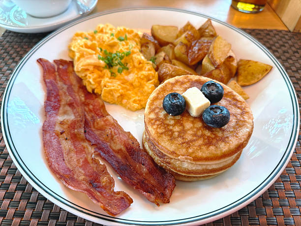 American breakfast with scrambled eggs, bacon, pancakes with blueberries, and roasted potatoes. Morning meal concept.
