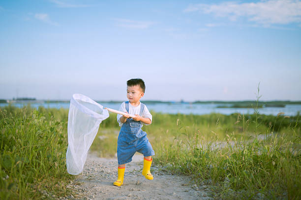 A cheerful young Asian boy dressed in blue overalls and yellow boots holds a white butterfly net while standing on a grassy path by a lake, enjoying a sunny day outdoors. The bright blue sky and natural surroundings enhance the playful and adventurous atmosphere of the scene.