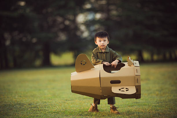 A young Asian boy stands on a grassy field in a park, playing with a handmade cardboard airplane toy. He wears casual green clothing and brown shoes, embodying childhood imagination and playful adventure in a natural outdoor setting.