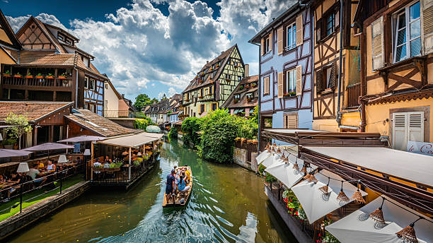 Little Venice (La Petite Venise) district in Colmar, Alsace, France. A small wooden boat carrying tourists glides along the tranquil canal, surrounded by colorful half-timbered houses and charming riverside cafés. The bright daylight and summer clouds highlight the romantic and fairy-tale atmosphere of an historic European town. Colmer in the Haut-Rhin department and Alsace region of north-eastern France.