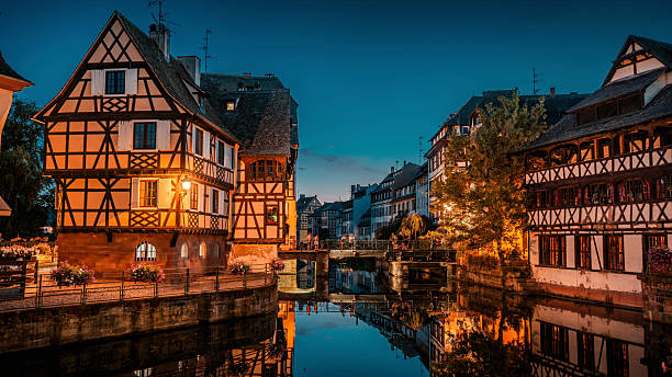 Twilight view of the picturesque La Petite France district in Strasbourg. Illuminated traditional half-timbered houses reflecting in the calm canal waters under a deep blue evening sky. Warm street lights, cobblestone walkways, and reflections in the water create a romantic and tranquil atmosphere. Strasbourg the largest city of the Grand Est region of eastern France, in the historic region of Alsace. Europe