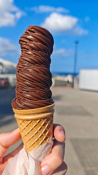 istock Hand holding a tall chocolate ice cream cone under a bright blue sky near the seaside 2242750907