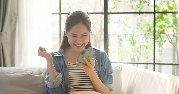 istock Asian woman smiling and using smartphone while relaxing on sofa in cozy home 2242751788