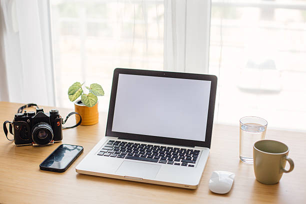 Workspace featuring a laptop with a blank screen, camera, smartphone, and drinks on a wooden desk
