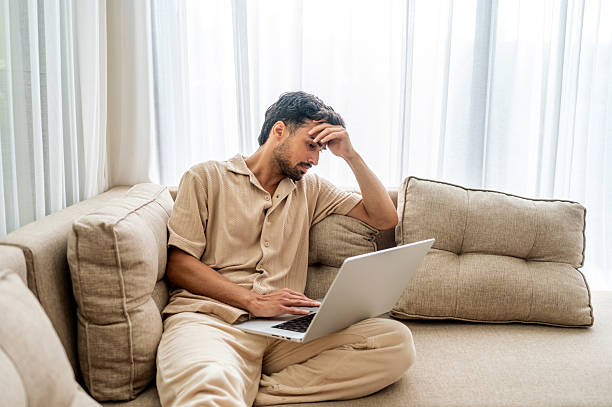 Contemplative Sad Man Working On Laptop On Couch In Bright Home Setting