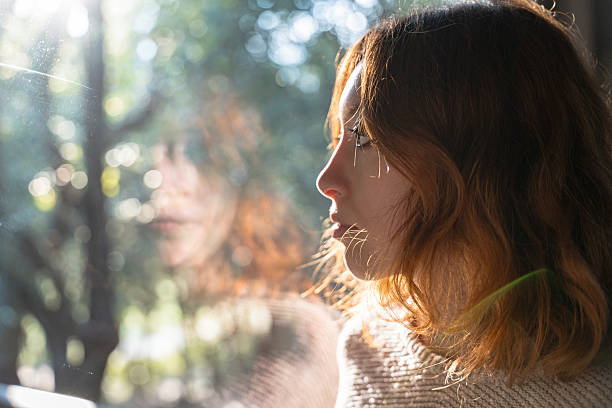 A young woman with auburn hair looks out the window as warm light streams in