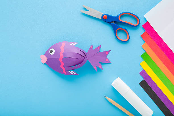 istock Purple fish shape, scissors, glue stick, pencil and colorful application paper on light blue table background. Pastel color. Closeup. Child handcraft. Top down view. 2242775881