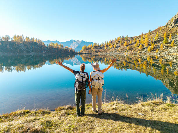 Two hikers with backpacks and hats standing by a calm mountain lake with clear reflections. Outdoor adventure and nature lovers appreciating scenic autumn landscape. Hiking and travel concept.