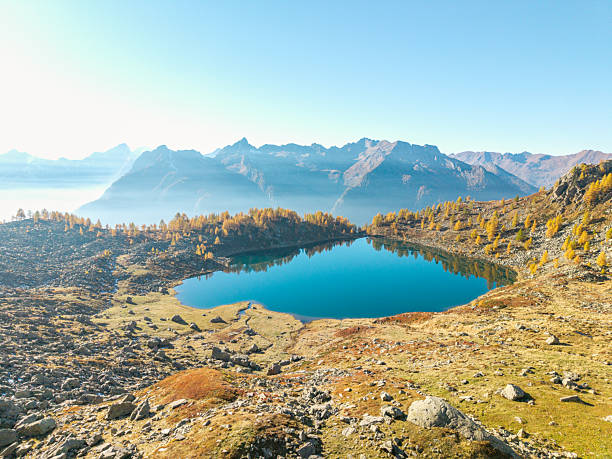 Clear blue mountain lake with golden autumn trees on rocky terrain. Scenic alpine landscape with distant mountain peaks under a bright sky. Nature and outdoor travel background.