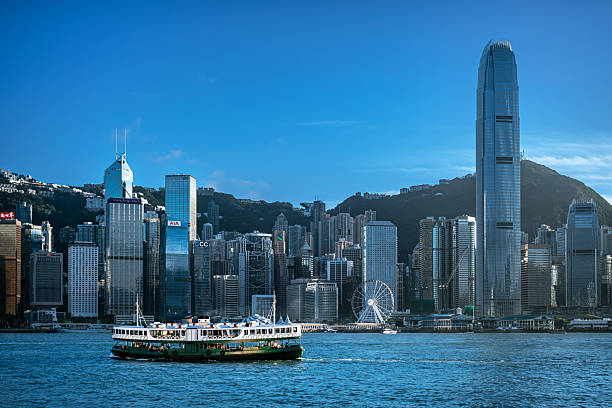 A vivid Hong Kong cityscape along Victoria Harbour featuring towering skyscrapers, glass façades, ferries, and a golden sunset glow over the skyline.